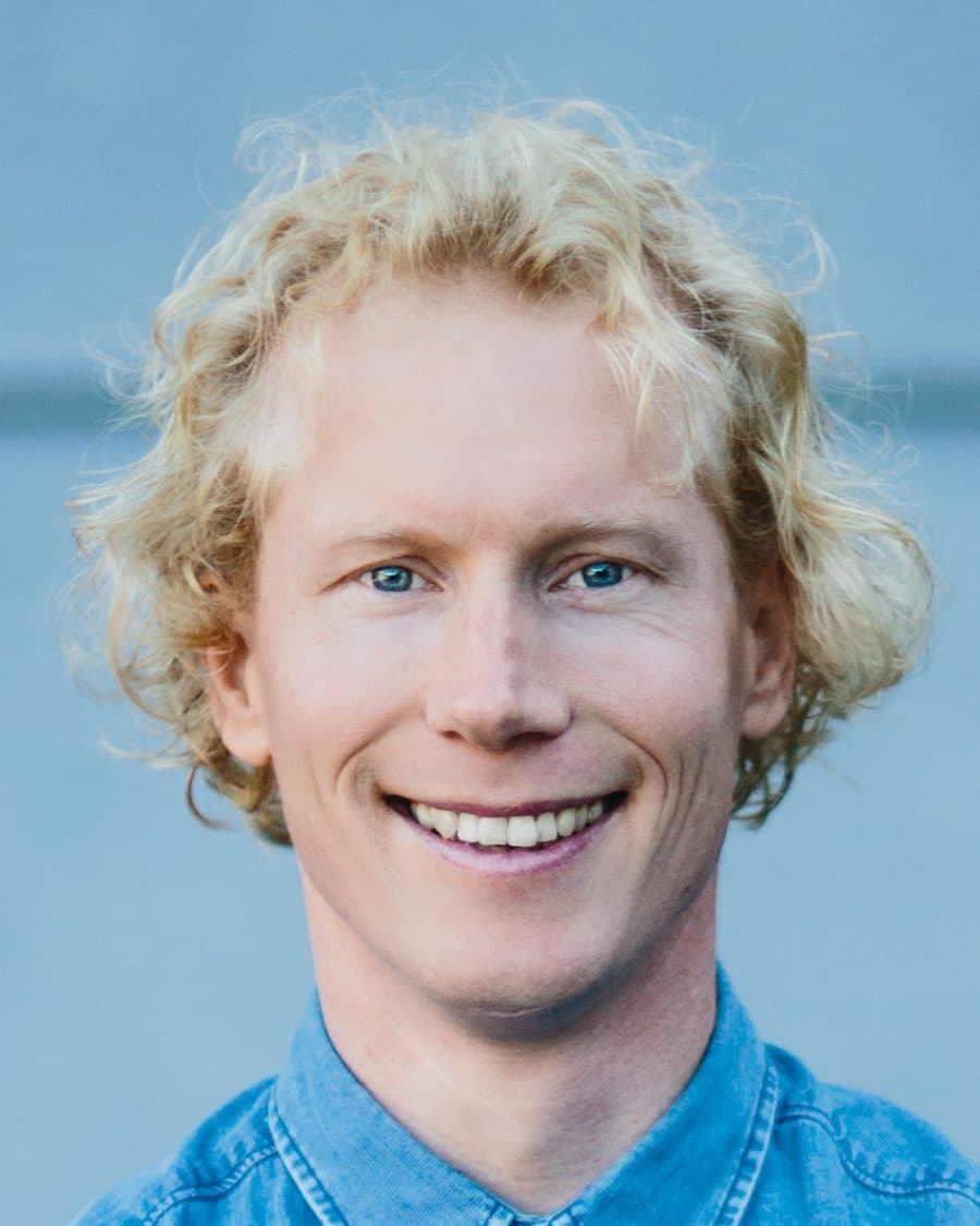 Aart Smit, StudieKeuzeCoach in Maastricht, lachend in een spijkerblouse voor een lichtblauwe muur