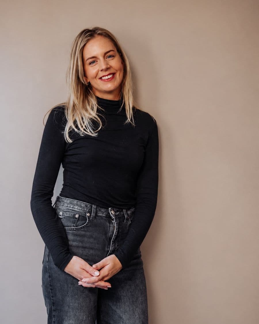 Aniek van der Hoeven, StudieKeuzeCoach in Nijmegen, lachend in een zwarte coltrui en een grijze spijkerbroek, met de handen gevouwen voor een taupe achterwand