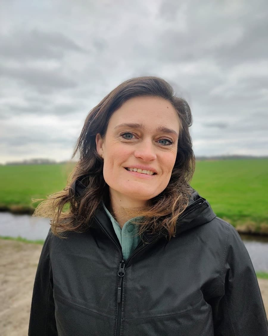 Jannemarie Kooijker, StudieKeuzeCoach in Zwolle, lachend in een donkere regenjas over een mintgroene fleece, buiten voor een groen weiland onder een bewolkte lucht