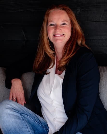 Angelina Muller, StudieKeuzeCoach in Rotterdam, lachend in een donkerblauw jasje en witte blouse op een grijze bank voor een zwarte houten wand