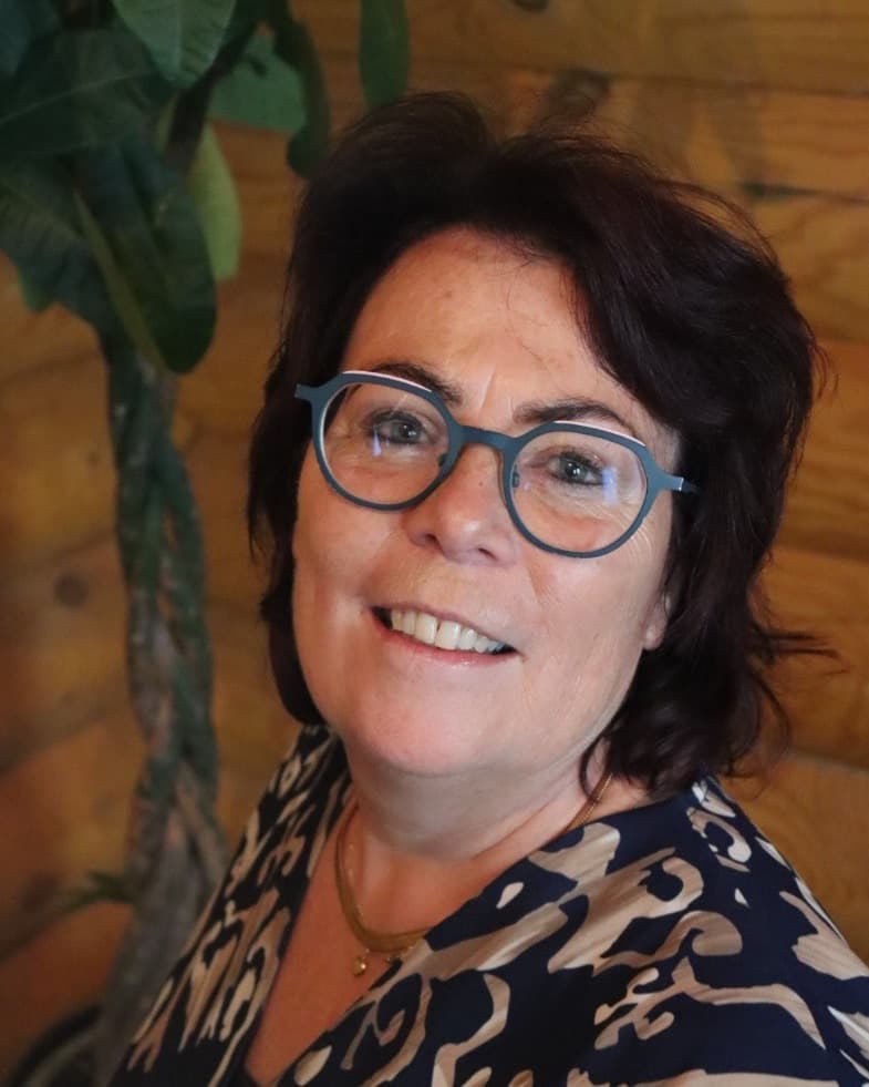 Ansje Swennenhuis, StudieKeuzeCoach in Twente, lachend met een blauwe bril en kort donker haar, in een donkerblauwe blouse met patroon voor een houten wand en een plant