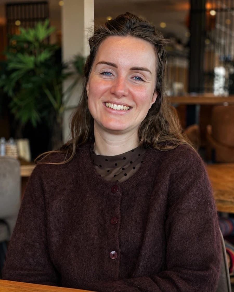 Astrid Raspe, StudieKeuzeCoach in Amersfoort, lachend in een bordeauxrood vest over een blouse met stippen, aan een houten tafel in een café